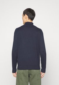 Man som står vänd bort med en mörk marinblå långärmad tröja och olivgröna byxor mot en enkel ljus bakgrund.