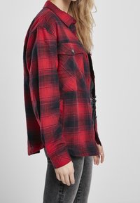 Kvinde iført en rød og sort ternet skjorte med brystlommer og slidte sorte jeans, stående sidelæns mod en ensfarvet baggrund.