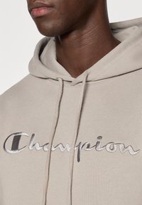 Μπεζ φούτερ με κουκούλα από βαμβακερό μείγμα, με εμφανές ασημένιο κέντημα "Champion" στο στήθος και κουκούλα με κορδόνι.