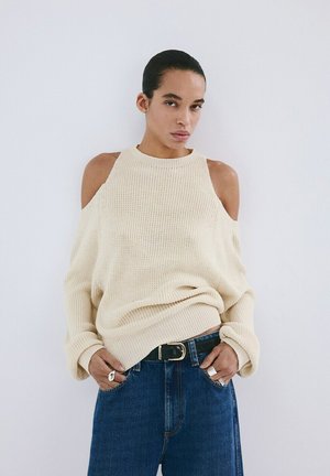 Personne aux cheveux tirés en arrière portant un pull en tricot beige à épaules dénudées et un jean bleu, les mains dans les poches, sur un fond uni.