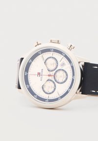 Στρογγυλό ασημένιο ρολόι χρονογράφου με λευκό καντράν, μπλε λεπτομέρειες, τρεις υποκαντράν, παράθυρο ημερομηνίας, κόκκινο δευτερόλεπτο δείκτη και μαύρο δερμάτινο λουρί.