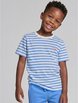Ein lächelnder Junge trägt ein blau-weiß gestreiftes T-Shirt und blaue Hose und steht vor einem einfarbigen hellgrauen Hintergrund.