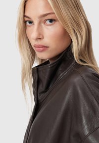 Brūna ādas jaka ar augstu apkakli, gludu tekstūru un elegantu dizainu. Iezīmes ietver redzamās šuves un pieguļošu siluetu.