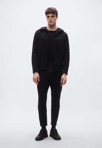 Czarna bluza z kapturem zapinana na zamek, o miękkiej fakturze, noszona na dopasowanej czarnej koszulce i dopasowanych zwężanych spodniach. Całość dopełniają czarne trampki.