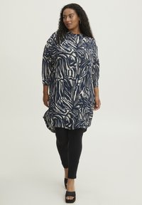 Marineblå tunika med abstrakt kremfarvet mønster, ¾ ærmer, asymmetrisk kant, parret med sorte leggings og kraftige sorte platformhæle.