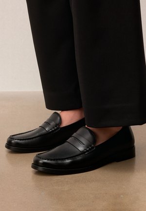 Zwarte leren loafers gedragen met zwarte cropped broeken op een beige vloer, met nadruk op schoen details en stiksels.