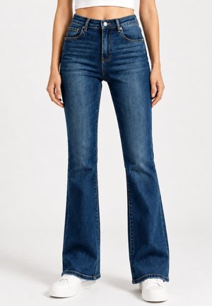 Donna che indossa jeans a zampa a vita alta blu scuro con sneakers bianche, in piedi davanti a uno sfondo chiaro e uniforme.