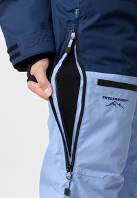 Blaue wasserdichte Skijacke mit schwarzen Akzenten und einem großen seitlichen Reißverschluss. Verfügt über ein Logo und eine Klettverschluss-Dichtung am Ärmel.