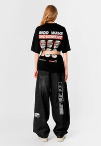 Camiseta gráfica negra con texto "MOVIMIENTO MOD WAVE", con ilustraciones de latas coloridas. Combinada con pantalones de mezclilla negros de pierna ancha.