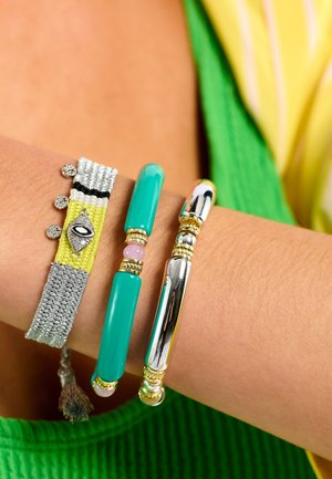 Trois bracelets sur un poignet : un bracelet tissé métallique gris texturé et jaune, un bracelet de perles turquoise lisses, et un bracelet argenté poli en forme de barre.