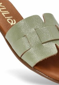 Sandalia plana de cuero texturizado verde con tiras entrelazadas y plantilla marrón. Cuenta con una suela plana y una marca decorativa en dorado.