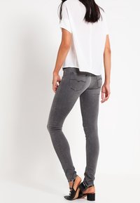 Jeans ajustados grises con una textura suave, diseño de cinco bolsillos, costuras sutiles y una cintura de altura media, combinados con una blusa blanca y tacones negros.