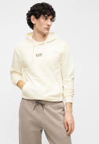 Jasnobeżowa bluza z kapturem z sznurkiem i przednią kieszenią z małym brązowym logo. Do tego jasnoszare spodnie dresowe.