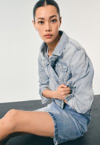 Lyseblå denimjakke med knaplukning, båret over en hvid top, parret med en matchende blå denimnederdel med frynsete kanter.