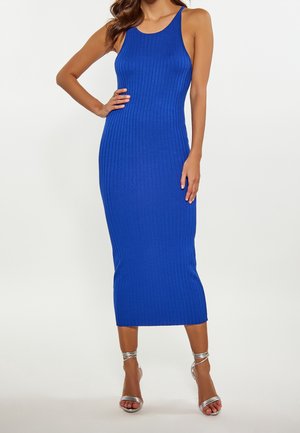Femme portant une robe midi bleu roi côtelée sans manches, associée à des sandales argentées à talons hauts et à bride à la cheville, posant avec une main sur la hanche.