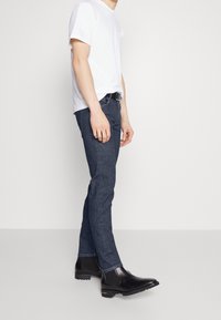 Svarta ankelskor, mörkblå jeans och en vit t-shirt. Elegant design med en mjuk yta och kontrasterande detaljer på bältet.
