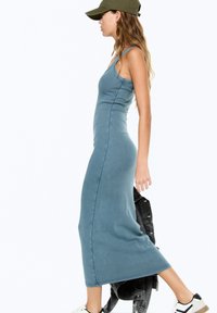 Rochie maxi din material teal, cu un design ajustat, bretele subțiri și cusături laterale. Asortată cu o jachetă din denim negru și adidași albi.