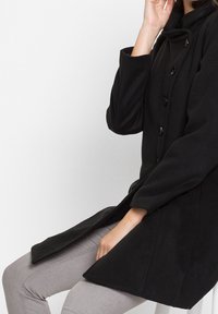 Svart långärmad kappa med hög krage, med knäppning fram och rak passform. Tyget verkar mjukt med en slät yta.