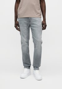 Ανοιχτό γκρι τζιν με ίσια γραμμή, διαθέτει πέντε τσέπες και διακριτική ξεθώριασμα. Συνδυάζεται με λευκά αθλητικά παπούτσια και μπεζ μπλούζα.