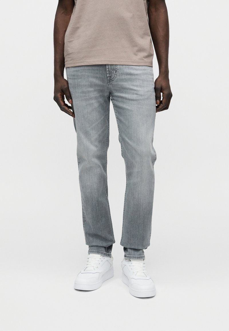 Ανοιχτό γκρι τζιν με ίσια γραμμή, διαθέτει πέντε τσέπες και διακριτική ξεθώριασμα. Συνδυάζεται με λευκά αθλητικά παπούτσια και μπεζ μπλούζα.