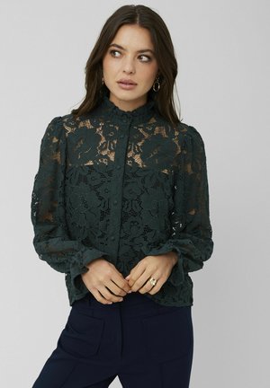Blouse en dentelle vert foncé avec un col haut, fermeture par boutons et manches longues bouffantes. Présente des motifs floraux et des poignets froncés.