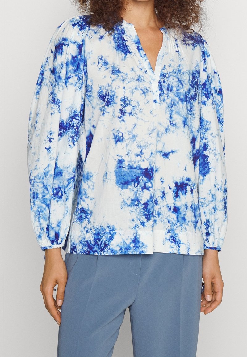 Femme portant un chemisier blanc avec un motif tie-dye bleu et des manches longues bouffantes, associé à un pantalon bleu.