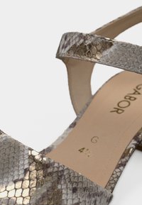 Πέδιλο με σχέδιο φιδιού και μεταλλικές λεπτομέρειες, με δύο λουράκια και μαξιλαρωτό πέλμα. Τα χρώματα περιλαμβάνουν ασημί, χρυσό και ταουπέ.