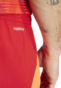 Rote Sportshorts verfügen über ein oranges Seitenpaneel und einen strukturierten Stoff. Der Markenname "AEROREADY" ist in weiß auf dem Bund aufgedruckt.