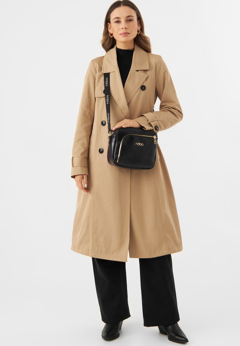 Trench coat beige con bottoni doppi, pantaloni neri e una borsa nera con hardware dorato e logo, indossata con stivali con tacco.
