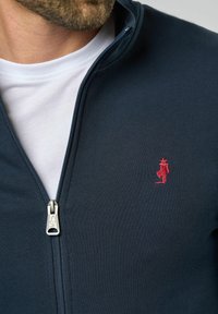 Marine blauwe fleece-jack met rits en een gladde textuur, voorzien van een witte onderhemd en een rode geborduurde logo-detail nabij de rits.