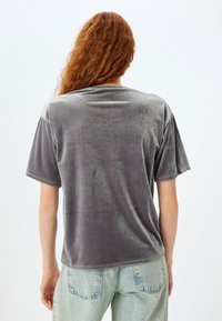 Camiseta de terciopelo gris con un corte relajado y mangas cortas. Presenta una textura suave y un brillo sutil. La vista trasera muestra un escote redondeado y detalles en las costuras.