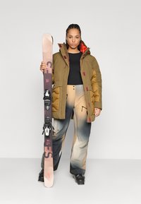 Stilfuld pufferjakke i oliven med maroon detaljer, sort top og gradient tan skibukser, holder pink ski med sorte bindinger.