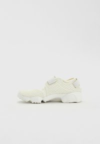 White athletic shoes with a textured mesh upper, featuring a strap for secure fit and a cushioned sole for comfort. Low-profile design.
