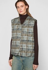 Padded vest w kolorze brązowym i szarym w kratę, z dekoltem w kształcie litery V, zapięciem na zatrzask i otworami na ręce. Noszony na ciemnym golfie.