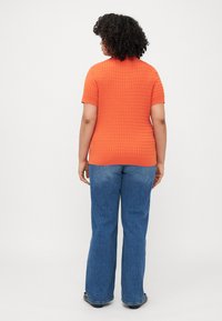 Oranje gebreid T-shirt met korte mouwen en een gestructureerd patroon, gecombineerd met blauwe flared jeans en zwarte schoeisel, gezien van achteren.