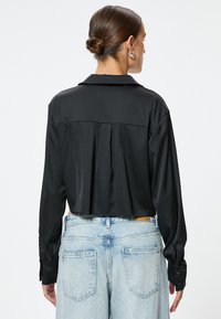Camisa de satén negra con cuello, mangas largas y pliegues en la parte posterior, combinada con pantalones de mezclilla de cintura alta en azul claro.