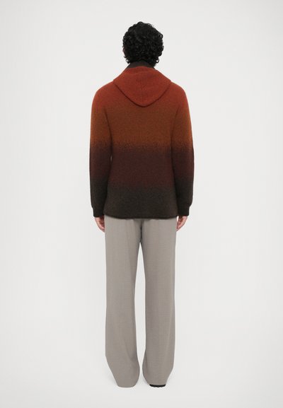 Sweater com capuz em um degradê de laranja a castanho-escuro, feito de material suave, com mangas longas e um ajuste relaxado, usado com calças cinza-claro.