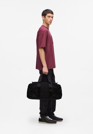 Black duffel bag with sturdy handles, reflective accents, two side pockets, and a textured fabric. Worn by a model in a maroon t-shirt.