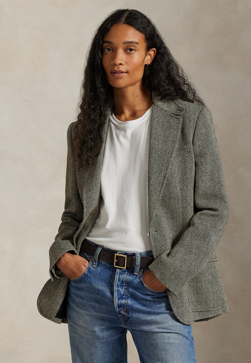 Vrouw met lang krullend haar die een visgraatblazer, wit T-shirt, blauwe spijkerbroek en zwarte riem draagt, staande met handen in de zakken.