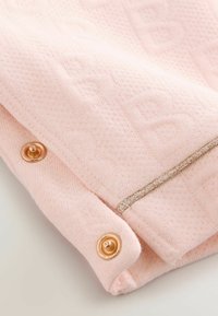 Vaaleanpunainen kangas, jossa on tekstuuripinta, kultaiseen sävyyn valmistetut napit ja koristeellinen hopea lanka reunan ympärillä.