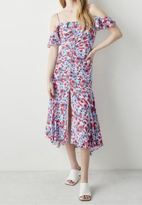 Robe midi florale avec des manches décolletées, fond bleu, fleurs rouges et vertes, devant froncé et fente centrale ; associée à des sandales blanches.