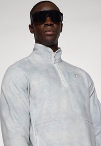 Heled hall fleece-kampsärg kõrge kaelusega, pool-zip ja reljeefne logo rinnal. Sile tekstuur ja minimalistlik disain.