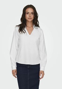 Blusa bianca con maniche lunghe, scollo a V e colletto ripiegato. Tessuto morbido e vestibilità leggermente ampia, abbinata a una gonna di jeans scura.