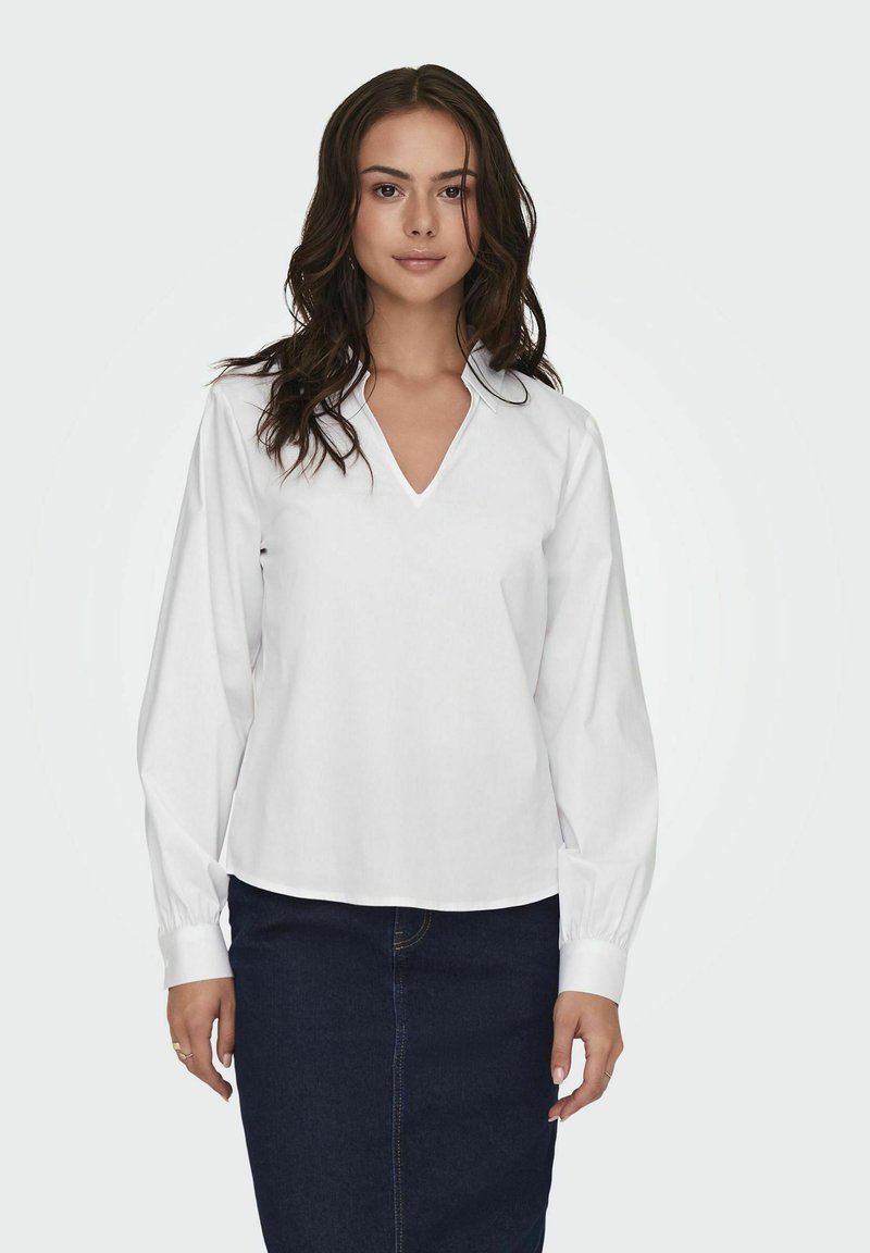 Blusa bianca con maniche lunghe, scollo a V e colletto ripiegato. Tessuto morbido e vestibilità leggermente ampia, abbinata a una gonna di jeans scura.