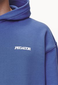 Sudadera azul con un logo blanco de "PEGADOR" en el pecho, mostrando el hombro y parte del cuello sobre un fondo liso.
