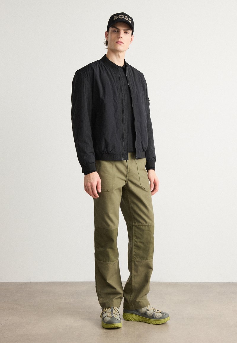 Czarna kurtka bomberka z zapięciem na zamek, oliwkowe spodnie cargo i szare buty z zielonymi akcentami, połączone z czapką w kolorze czarnym z złotym napisem.