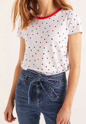 Frau trägt weißes kurzärmeliges T-Shirt mit mehrfarbigen Tupfen und rotem Halsausschnitt, kombiniert mit hoch taillierten blauen Jeans, gebunden mit einem Jeansgürtel.