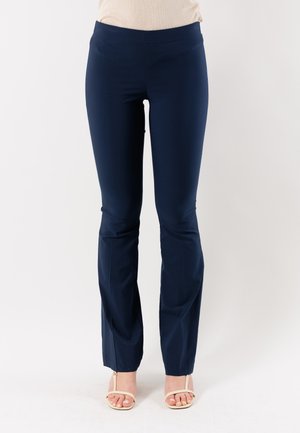 Donna in pantaloni aderenti blu navy e sandali beige con punta aperta che sta in piedi davanti a uno sfondo bianco semplice.