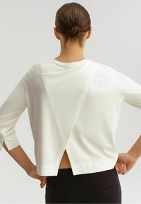 Top bianco in tessuto morbido con una vestibilità ampia, caratterizzato da uno spacco profondo sulla schiena e maniche a tre quarti. Design minimalista con linee pulite.