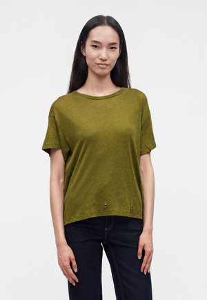 Femme aux longs cheveux foncés portant un t-shirt vert olive à manches courtes et un jean foncé, debout devant un fond uni clair.